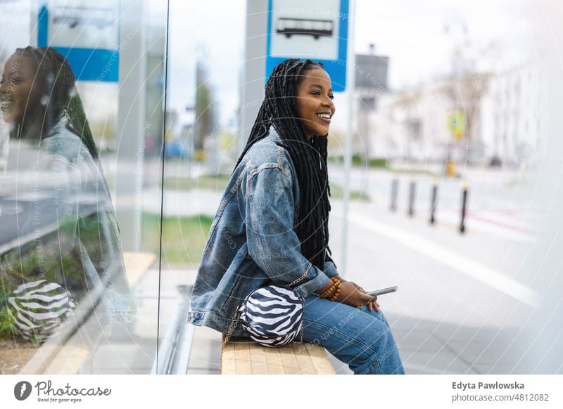 Junge Frau, die an einer Bushaltestelle auf einen Bus wartet cool sorgenfrei selbstbewusst Tag Jahrtausende Rastalocken genießen authentisch positiv