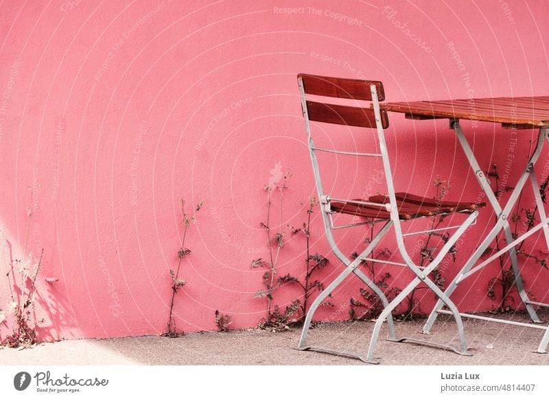 Tisch und Stuhl eines Straßencafés, an der rosa gestrichenen Hauswand kämpfen verdorrende Gewächse um Licht und Wasser, die Gäste lassen auf sich warten. Stadt