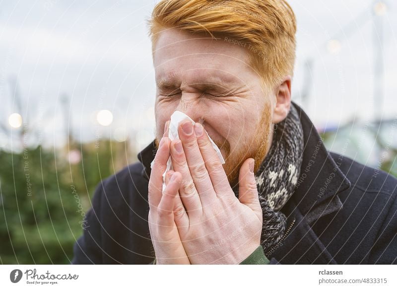 Mann niest und schnäuzt seine Nase in ein Taschentuch im Winter mit Allergie oder eine Infektion ein Virus. Porträt eines rothaarigen Mannes im Freien. sinus
