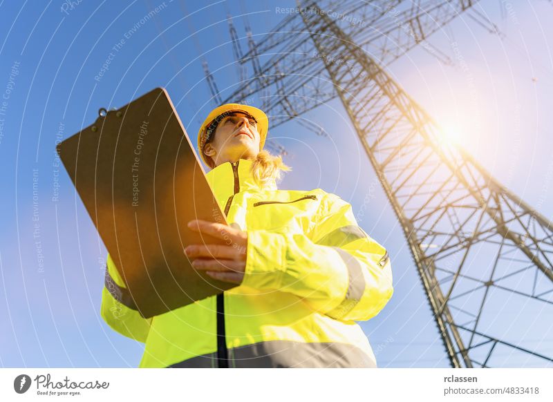 Eine Elektroingenieurin steht mit einem Klemmbrett in einem Elektrizitätswerk und beobachtet die Planungsarbeiten für die Stromerzeugung an Hochspannungsmasten.