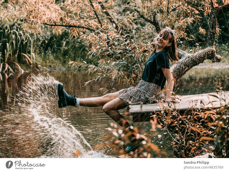 Junge Frau sitzt auf einer Holzbrücke und spritzt mit den Stiefeln Wasser auf Brücke nass Sommer Außenaufnahme Farbfoto feminin Freude stylisch fröhlich