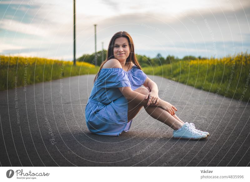 Candid Porträt eines schüchternen jungen Mädchens in einem schönen blauen Sommerkleid mit weißen Turnschuhen sitzen auf der Straße mit einem natürlichen atemberaubenden Lächeln bei Sonnenuntergang. Mode-Stil. Europäisches Modell