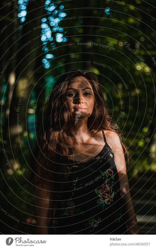 Nachdenklicher Blick einer 24-jährigen Frau mit dunkelbraunem Haar in einem Wald bei Sonnenuntergang. Unverfälschtes Porträt einer hübschen Frau im Sommerkleid. Versteckt in den Schatten