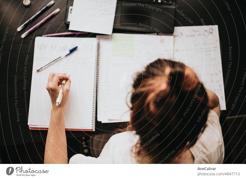 Draufsicht auf den Rücken einer jungen Frau, die zu Hause am Schreibtisch studiert und arbeitet, Hausaufgaben macht, sich mit dem Lehrbuch auf die Prüfung vorbereitet und Notizen macht, selektiver Fokus auf den Bleistift.