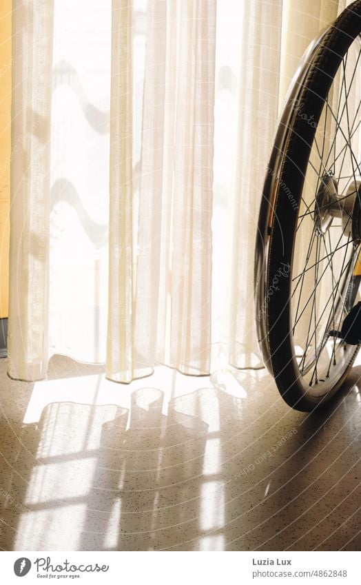 Am Fenster: angeschnitten zu sehen das Rad eines Rollstuhls. Sonnenlicht fällt durch die zarten, hellen Gardinen und wirft Schatten auf den Boden. sonnig