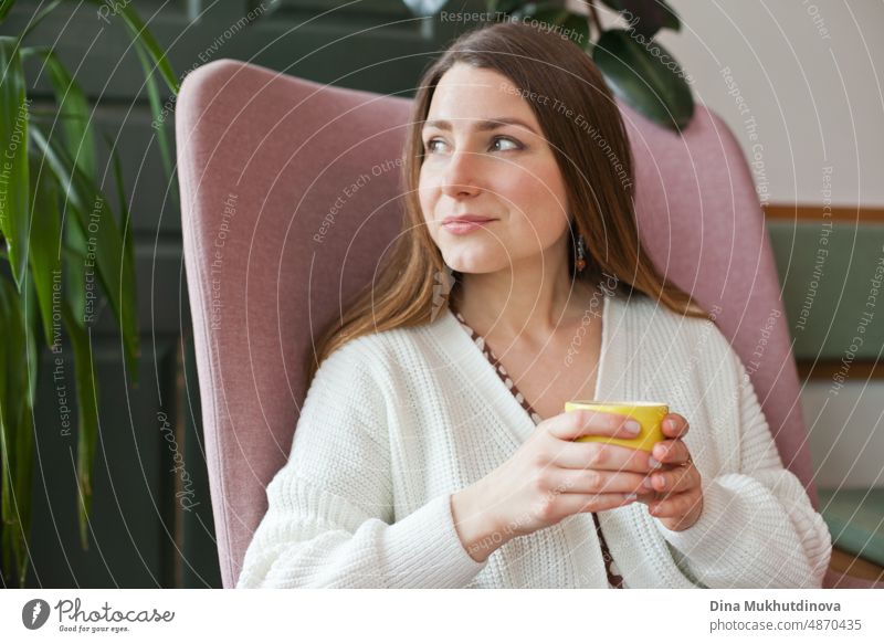 Schöne Frau trinkt Kaffee im Café sitzt in rosa Stuhl. Candid Millennial Lebensstil. Attraktives Gesicht einer erwachsenen Frau in ihren Dreißigern. Blick aus dem Fenster im Restaurant, hält eine Tasse Kaffee. Kaffeepause in der Cafeteria bei der Arbeit im modernen Büro.