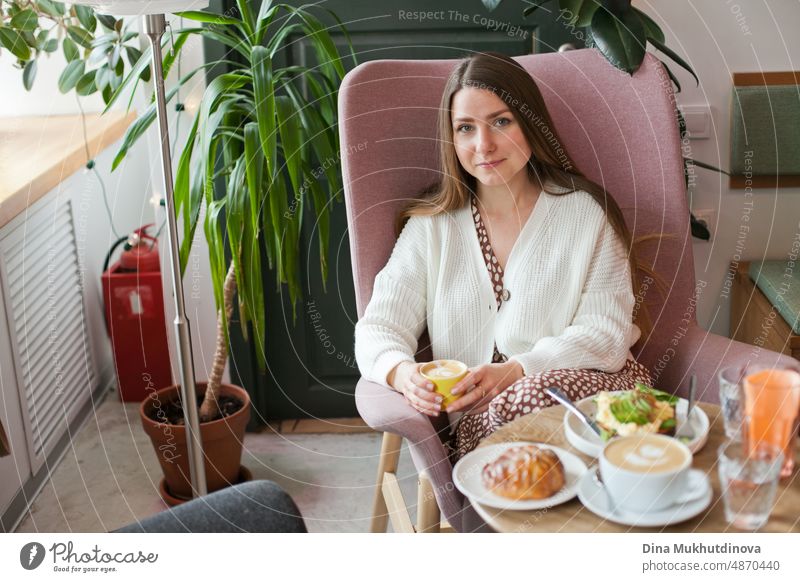Schöne Frau trinkt Kaffee im Café sitzt in rosa Stuhl. Candid Millennial Lebensstil. Attraktives Gesicht einer erwachsenen Frau in ihren Dreißigern. Blick aus dem Fenster im Restaurant, hält eine Tasse Kaffee. Kaffeepause in der Cafeteria bei der Arbeit im modernen Büro.