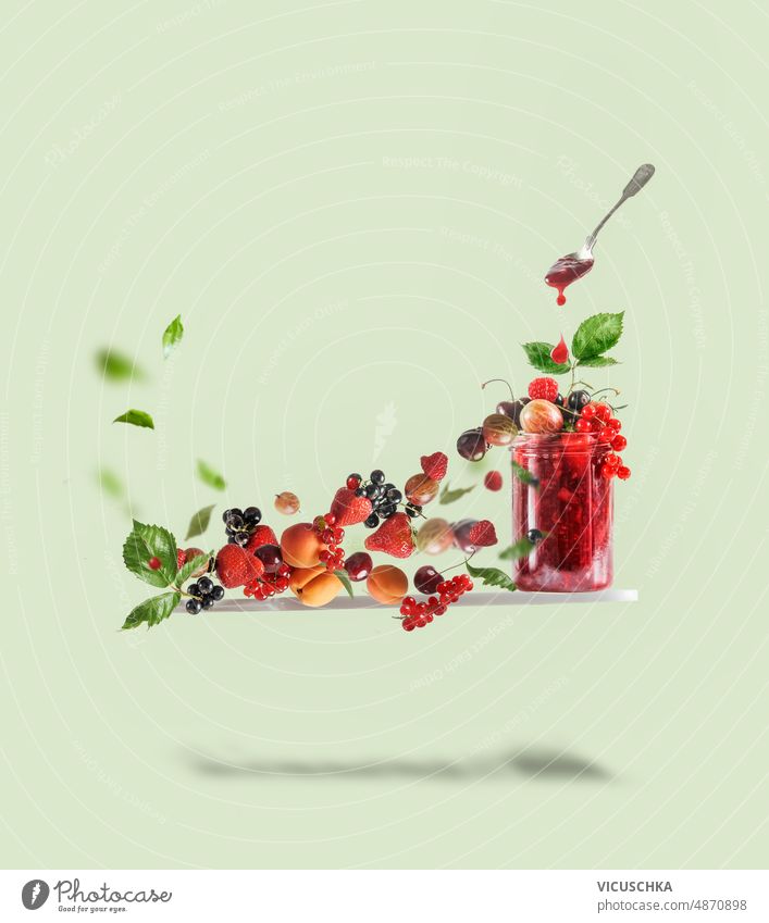 Fliegendes Schneidebrett mit rotem Marmeladenglas, Löffel und verschiedenen Beeren auf blassgrünem Hintergrund. Kreatives Essen fliegen Glas Johannisbeeren