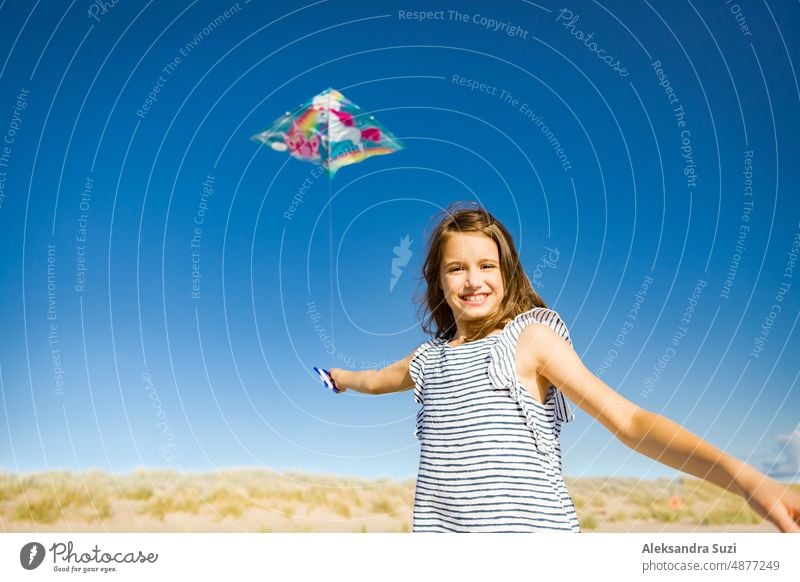 Nettes glückliches kleines Mädchen im Sommerkleid läuft mit fliegenden Drachen auf leeren Sandstrand. Schöner sonniger Tag, blauer Himmel. aktiv Aktivität