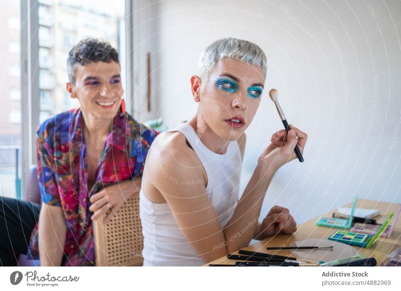 Mann schaut auf seinen Freund, der sich am Frisiertisch schminkt schwul anwendend Make-up Spiegel Dressing Tisch Lächeln Paar Transgender Lidschatten Waschtisch