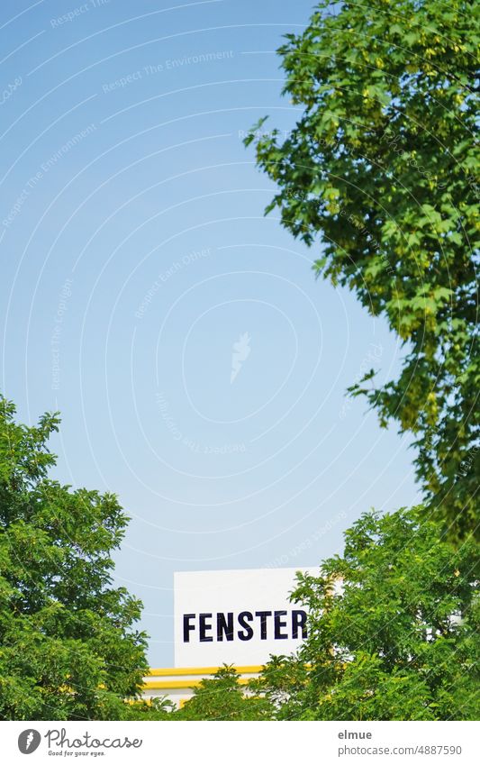 FENSTER steht in Großbuchstaben an einer Werbetafel hinter grünen Laubbäumen / Durchblick Fenster Gebäude Ausblick Bäume Werksgelände Werbung Himmel himmelsblau