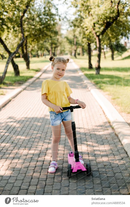 Ein aktives kleines Mädchen fährt an einem Sommertag auf einem Pfad in einem Park im Freien mit einem Motorroller. Aktiver Sport für Kinder in der Saison. Gesunder Lebensstil in der Kindheit