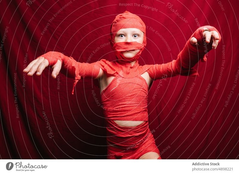 Niedliches kleines Kind im Mumienkostüm, das im roten Studio schreit und gestikuliert Halloween Tracht Junge beängstigend Porträt spukhaft sich[Akk] melden