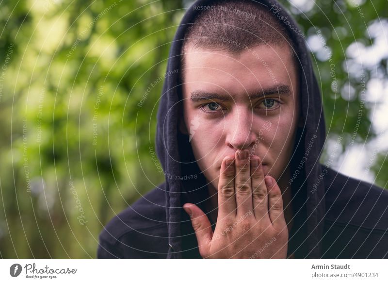 Porträt eines jungen ernsten Mannes mit Kapuzenpulli und Hand vor dem Mund deckend stumm Finger Aussehen Teenager Blick männlich schön lässig Kaukasier