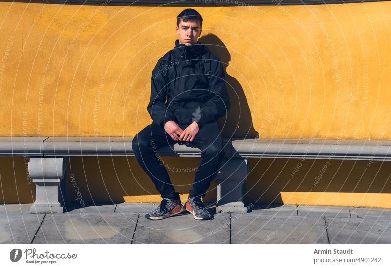 junger Mann sitzt auf einer Steinbank mit gelber Wand im Hintergrund Sitzen Bank ernst entspannt Pause Rucksack Reise Porträt Teenager Blick männlich schön