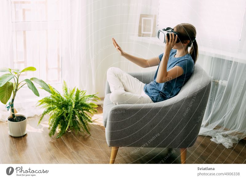 Das Mädchen sitzt mit einer virtuellen Brille auf einem Stuhl und lernt online oder spielt virtuelle Spiele. Moderne Technologien, virtuelles Lernen, moderne Freizeit und Bildung