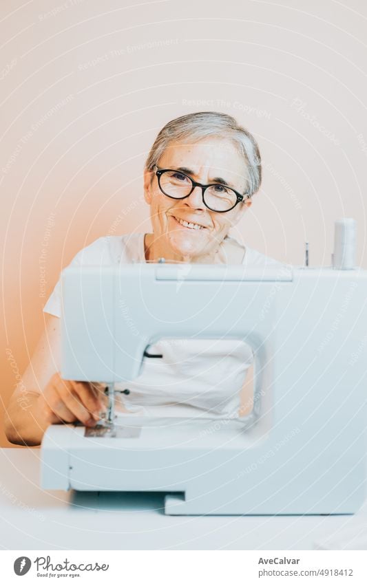 Alte Frau Schneiderin arbeitet an Nähmaschine lächelnd glücklich zu Kamera während nähen. Wiederverwendung alter Stoffe für neue Kleidung, Nachhaltigkeit. Alte Menschen neues Hobby zu Hause, Senioren geistige Aktivität und Arbeit
