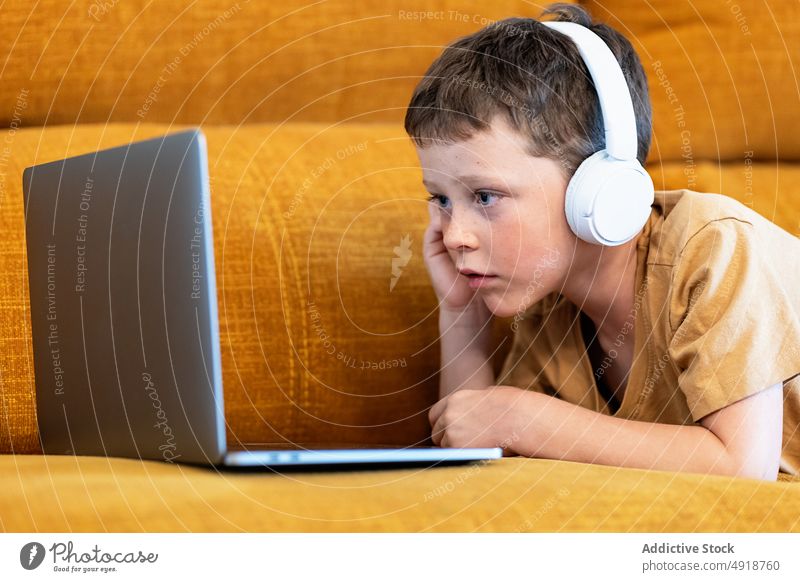 Junge sitzt auf einem Sofa mit Kopfhörern auf dem Kopf und benutzt den Laptop Kind Computer Kaukasier Technik & Technologie männlich ernst Konzentration