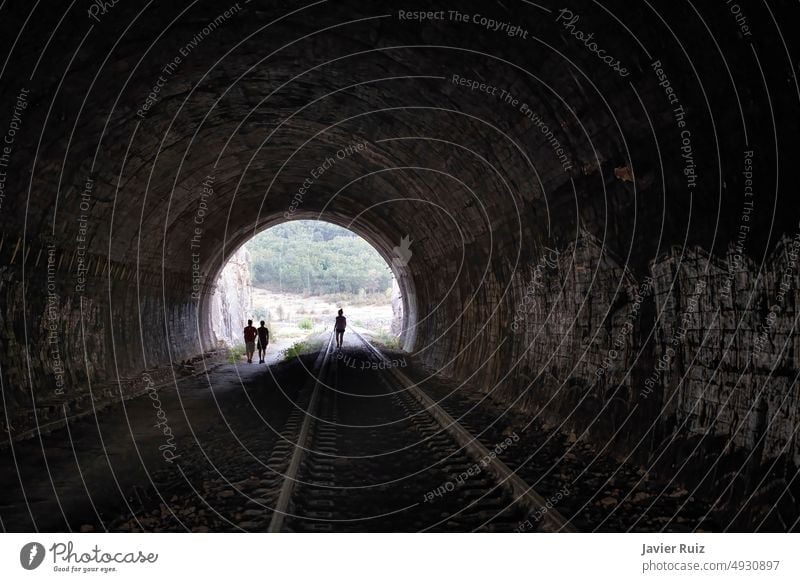 der Ausgang eines alten, dunklen Eisenbahntunnels, von innen gesehen, mit der Silhouette von drei Menschen, die in der Ferne aus dem Tunnel kommen, Konzept der Überwindung