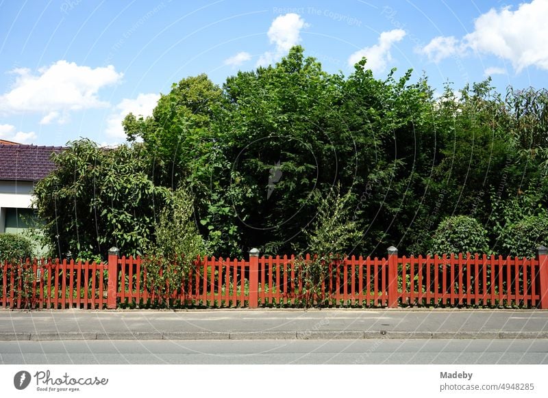 Schöner roter Holzzaun im Sonnenschein vor einem grünen Garten im Sommer mit blauem Himmel und weißen Wolken in Oerlinghausen bei Bielefeld am Hermannsweg im Teutoburger Wald in Ostwestfalen-Lippe