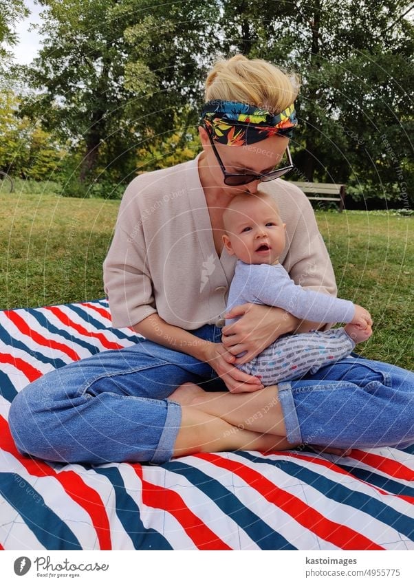 Moderne trendy Mutter küssen und streicheln sein Baby Junge Kind auf einer Matte in einer Wiese im Freien in der öffentlichen Stadtpark Mama Mutterschaft