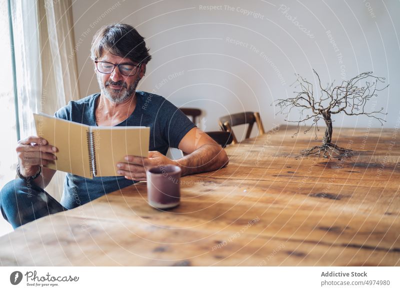 Konzentrierter älterer Mann, der zu Hause sein Notizbuch liest lesen Notebook fokussiert Windstille Inhalt Konzentration sitzen Tisch hölzern Tasse Überprüfung