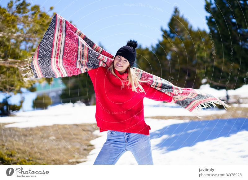 Glückliche junge blonde Frau, die in einem Wald in den verschneiten Bergen ihren Schal im Wind schwenkt. Winter Mädchen Schnee Freude Lächeln warm weiß heiter