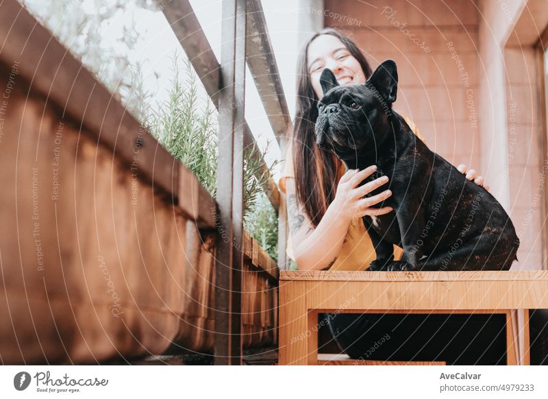 Junge Frau, die ihre schwarze französische Bulldogge streichelt und umarmt, während sie auf einer Holzbank sitzt. Der Hund beobachtet die Stadt und das Mädchen lächelt. Neues Leben in der Stadt, offener und spielerischer Platz auf der Terrasse zum Spielen und Entspannen