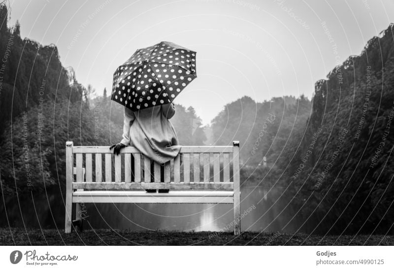 [HH Unnamed Road] Rückansicht einer Frau in einem hellen Mantel mit einem gepunkteten Regenschirm. Sie sitzt auf der Lehne einer Bank am Rande eines Sees.