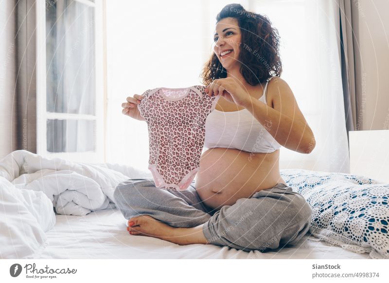 Glückliche schwangere Frau mit Babykleidung Kleidung Schwangerschaft Bauch Konzept heimwärts schön Mutter natürlich Licht Morgen Mutterschaft erwartend Bett
