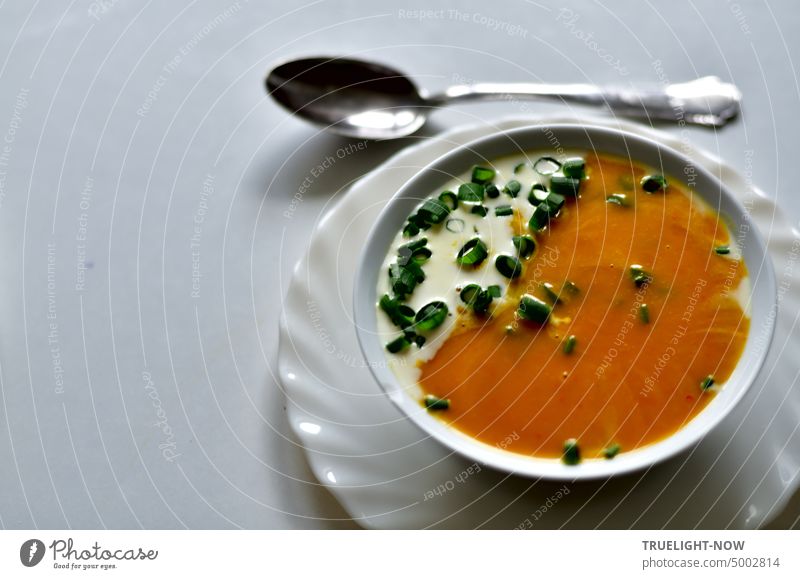 Kürbissuppe mit etwas Sahne und Lauchzwiebelgrün kleingeschnitten in weißer Schale auf weißem Teller und ein Suppenlöffel orange Schnittlauch Wärme Harmonie