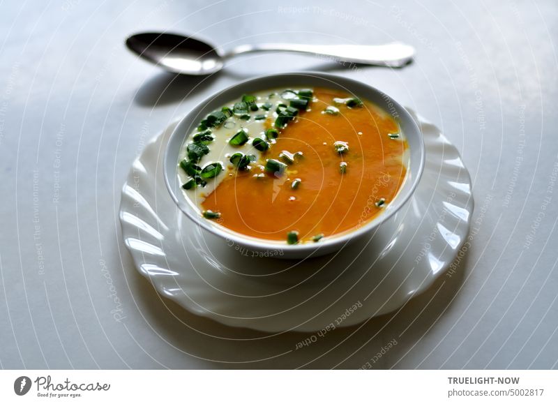 Kürbissuppe frisch püriert, dazu etwas Sahne und Lauchzwiebelgrün kleingeschnitten in weißer Schale auf weißem Teller und ein Suppenlöffel orange Schnittlauch