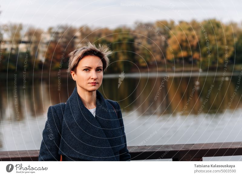 Eine junge Frau in einem Park im Herbst am Fluss schaut in die Kamera fallen Kurze Haare blond Selbstvertrauen Lebensstile Erwachsener Freizeit