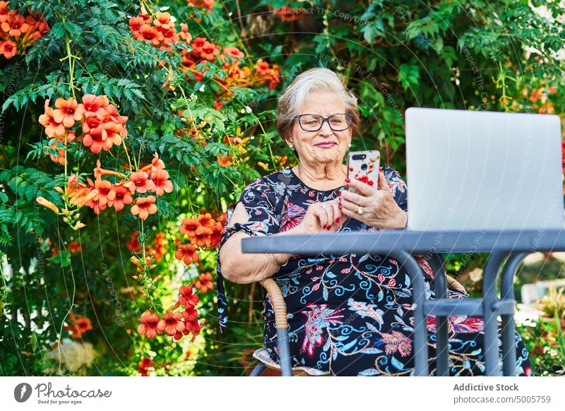 Ältere Dame surft mit Smartphone am Gartentisch mit Netbook Frau Telefon Tisch Surfen Hinterhof Stuhl gealtert älter Senior Blüte grün Park Mobile modern Sommer