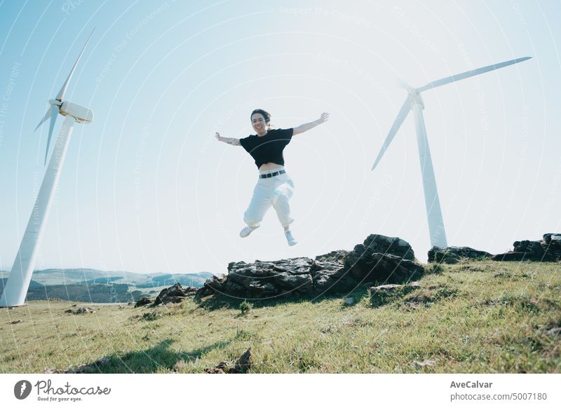 Junge Frau, die springt und Spaß hat und die Freiheit feiert, indem sie die Arme in den Himmel streckt und in die Kamera lächelt, umgeben von elektrischen Windrädern. Blasen Haare und entspannen, saubere Erde Zukunft Konzept