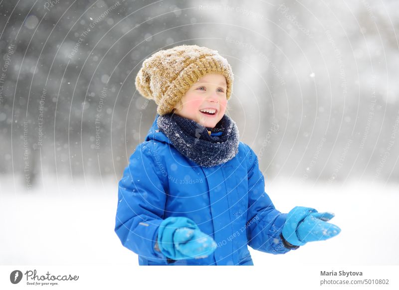 Kleiner Junge hat Spaß beim Spielen mit frischem Schnee während des Schneefalls. Baby fängt Schneeflocken auf Handschuhe. Kind in warmer Kleidung, Mütze, Handschuhe und Schal gekleidet. Aktive Winter im Freien Freizeit für Kind