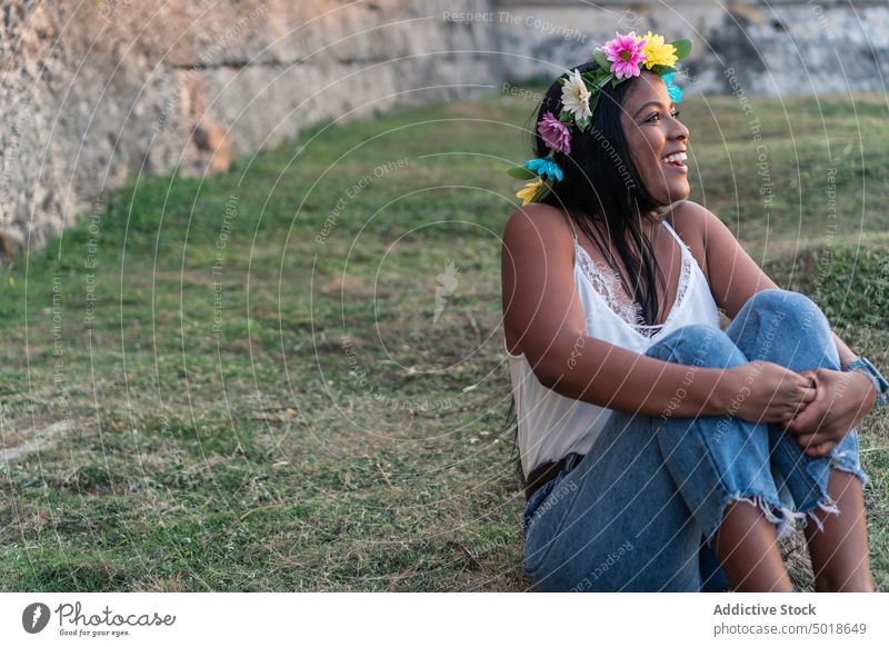 Lächelnde ethnische Frau mit Blumenkranz im Feld Totenkranz Gelassenheit romantisch charmant geblümt Knie umarmend sanft Natur Angebot Windstille Sommer feminin