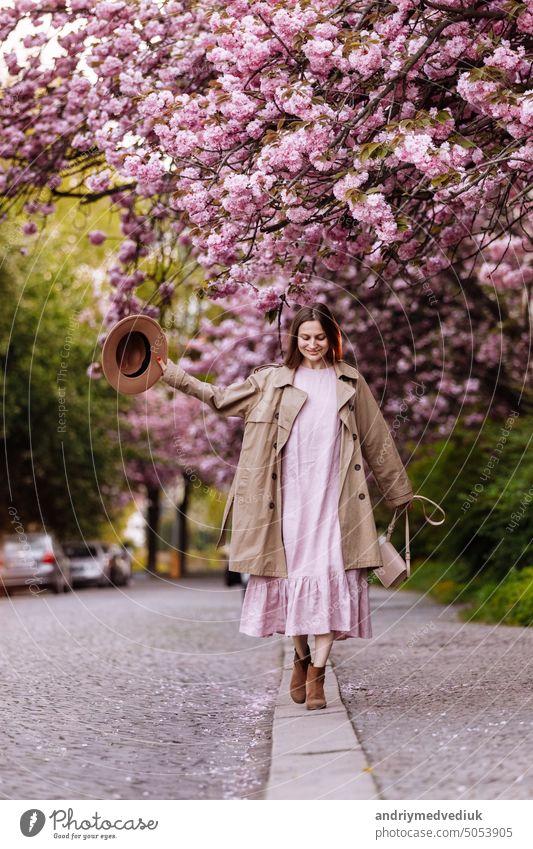 junge schöne stilvolle Frau in Hut und rosa Kleid zu Fuß in der Nähe von sakura Blumen im Park. Frühling Konzept. Uzhhorod, Ukraine Sakura Mode Schönheit