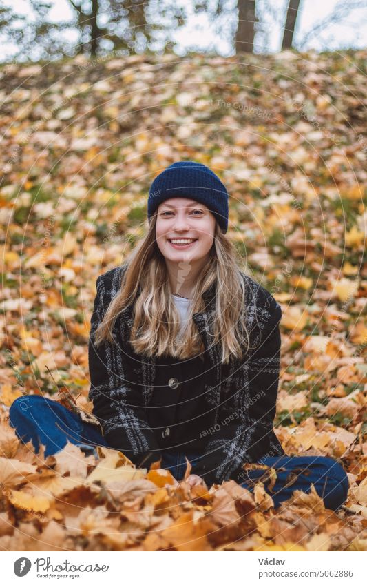 Lächelnde Brünette sitzt in einem Haufen bunter Blätter in einem Eichenwald und wirft Blätter. Candid Porträt einer jungen realen Frau im Herbst Kleidung in der Herbstsaison