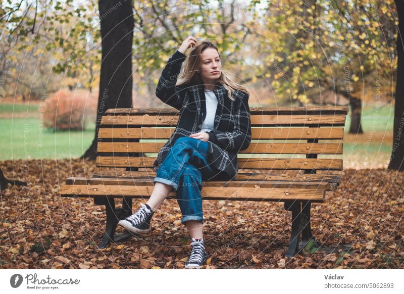 Lächelnde Brünette, die auf einer ornamentalen Bank in der Mitte des Parks sitzt und die Herbstfarben genießt. Unverhohlenes Porträt einer jungen echten Frau in Herbstkleidung