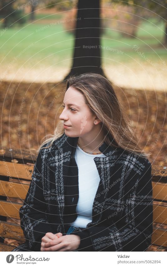 Lächelnde Brünette, die auf einer ornamentalen Bank in der Mitte des Parks sitzt und die Herbstfarben genießt. Unverhohlenes Porträt einer jungen echten Frau in Herbstkleidung