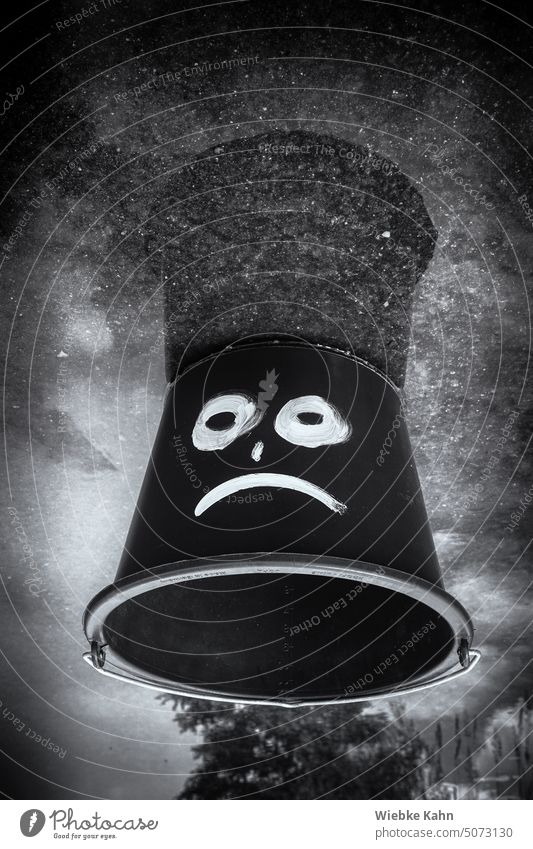 Schwarz-weiß Bild. Schwarzer Eimer mit weiß aufgemalten traurigen Gesicht in Pfütze. Pfütze Flüssigkeit Pfützenbild witzig Smiley Smiley-Symbol Schwarzweißfoto