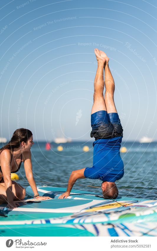 Mann macht Kopfstand beim Yoga, während Frau am Meer zusieht ausführen zuschauen Paddelbrett üben MEER Resort wolkenlos sirasana Blauer Himmel Gleichgewicht