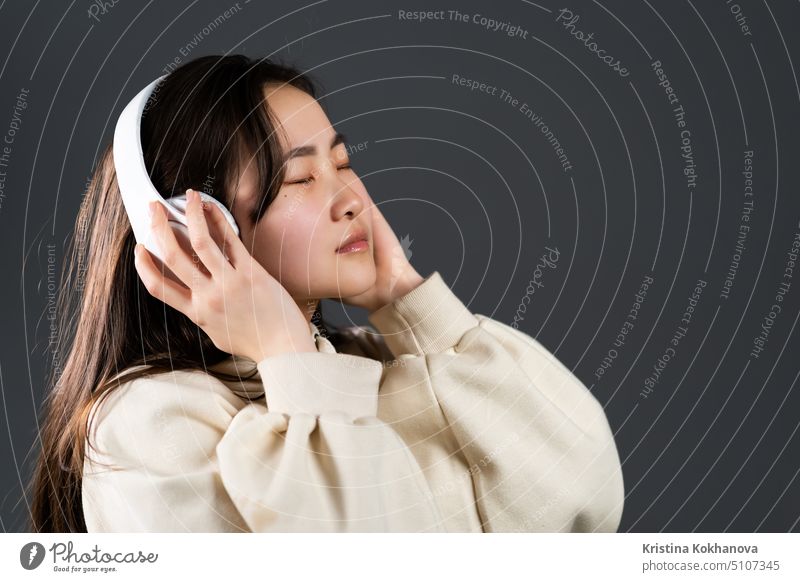 Netter asiatischer Teenager, der mit Kopfhörern Musik hört. Porträt von Millennial hübsches Mädchen mit langer Frisur auf grauem Hintergrund Licht Glamour Mode