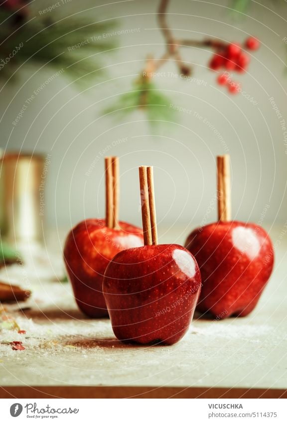 Rote Äpfel mit Zimtstangen auf Küchentisch mit Zucker an Wand Hintergrund mit Tannengrün. Zucker glasierte Äpfel. Leckere Weihnachtssüßigkeiten. Vorderansicht.
