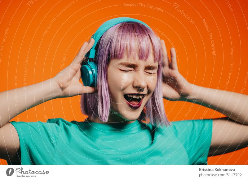 Außergewöhnliche Frau mit gefärbten violetten Haaren hört Musik mit Kopfhörern und singt auf orangefarbenem Hintergrund. Einzigartige Hipster Mädchen tanzen