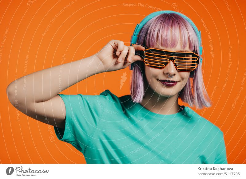 Außergewöhnliche Frau mit gefärbten violetten Haaren hört Musik mit Kopfhörern und singt auf orangefarbenem Hintergrund. Einzigartige Hipster Mädchen tanzen mit Gitter Sonnenbrille.