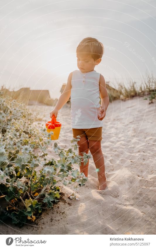 Süßer kleiner Junge gießt Pflanzen mit Gießkanne am Sandstrand. Sommer sonniger Tag. Kleinkind mit buntem Topf. Natürliche ästhetische Porträt des Kindes.