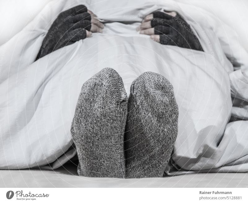 Eine Person liegt in einem Bett. Sie hat dicke Socken und Handschuhe an. Frau kalt Kälte frieren Winter Frost Bettwäsche Heizung Heizkosten teuer Energie