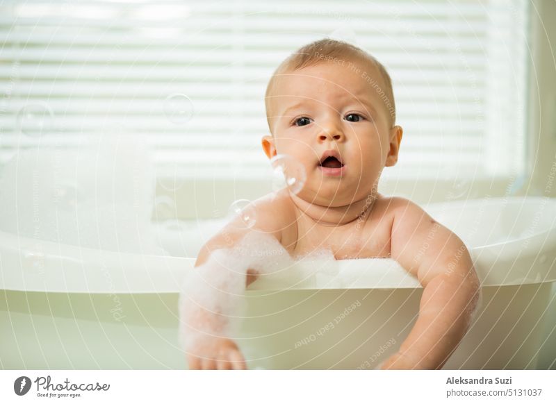 Nettes kleines Baby sitzt in der weißen Badewanne mit Schaum und Seifenblasen. Nimmt ein Bad und spielt mit Spielzeug. Baby-Hygiene. 6 Monate 6 Monate Baby